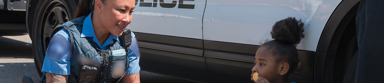 A female law enforcement officer talks with a child in front of a police cruiser.